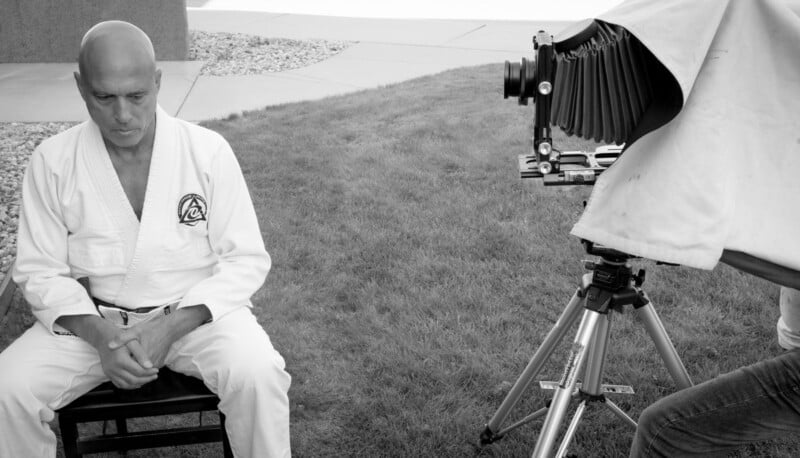 A man in a martial arts uniform sits on a chair outdoors, looking down pensively, while an old-fashioned camera on a tripod is positioned in front of him. The photo is in black and white.