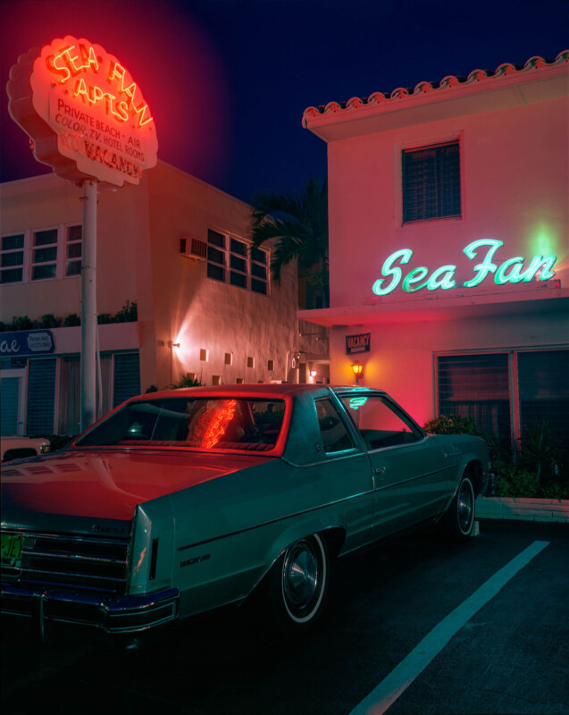 Un viejo automóvil turquesa estacionado fuera del apartamento del ventilador del mar, iluminado por letreros de neón e iluminación cálida. El edificio y el logotipo vintage crean una atmósfera nostálgica de hoja media.