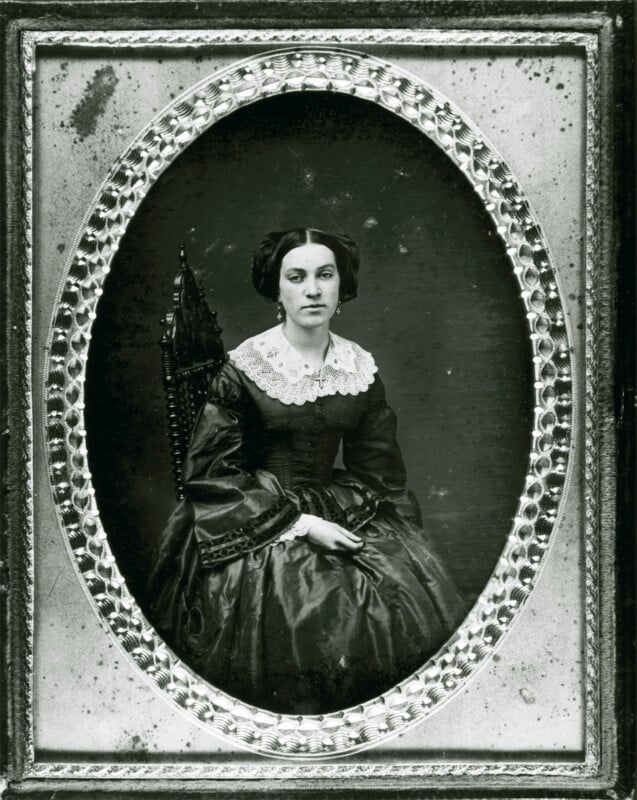 A vintage black-and-white portrait of a seated woman wearing a dark dress with a wide lace collar, her hair styled in an 1800s fashion, framed by an ornate oval border.