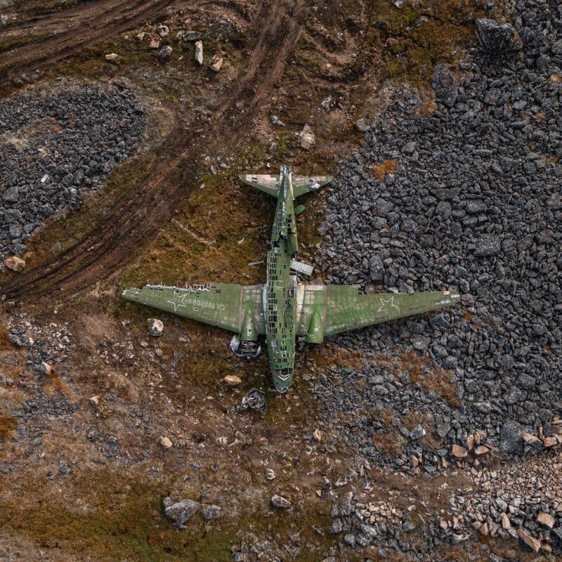 Aerial view of an abandoned, green military airplane wreck lying on rocky, barren terrain surrounded by dirt paths and scattered stones.