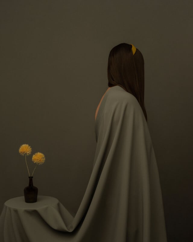 Junto a una pequeña mesa cubierta con un mantel gris estaba sentado un hombre, envuelto en un paño gris, de espaldas a la distancia. Hay un jarrón oscuro sobre la mesa con dos flores amarillas. El fondo es de suaves tonos grises y el personaje lleva una diadema amarilla.