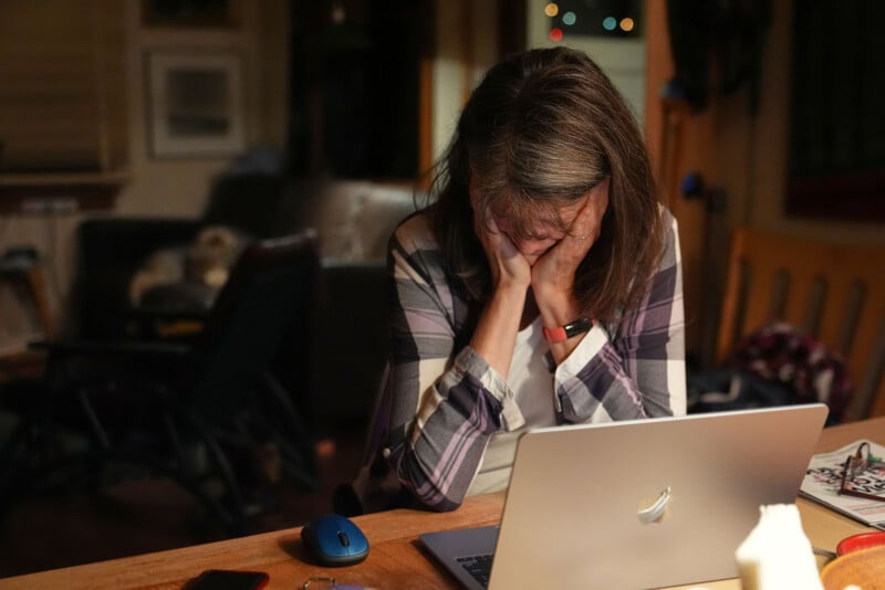 Una mujer sentada en una mesa con la cabeza entre las manos y parece nerviosa frente a una computadora portátil abierta. La habitación está poco iluminada y desordenada, lo que sugiere un sentimiento de abrumador o frustración.