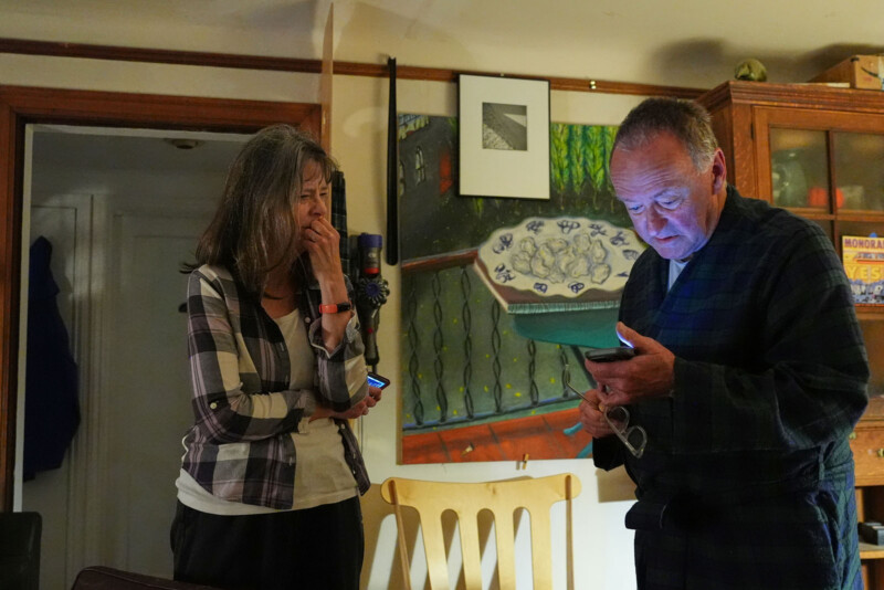 Una mujer se encuentra de pie con la mano tapándose la boca mientras un hombre en bata mira un teléfono inteligente, ambos en una habitación cálidamente iluminada con coloridas obras de arte y muebles de madera al fondo.