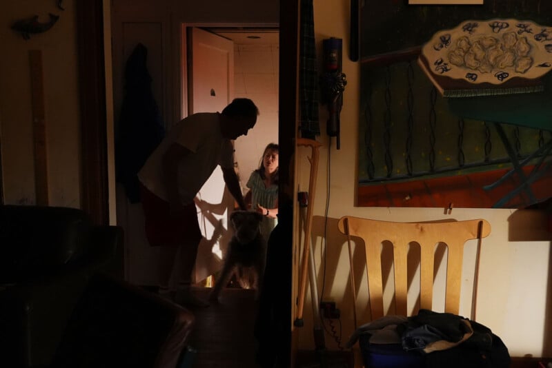 En una puerta con poca luz había un hombre y una niña con un perro. El hombre intenta alcanzar al perro y la cálida luz de otra habitación ilumina suavemente la escena. En primer plano se puede ver un cuadro y sillas de madera.