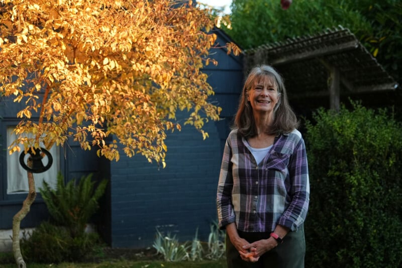 Una mujer de pelo largo y gris está parada en el jardín sonriendo, vestida con una camisa a cuadros y un top blanco. Detrás de ella hay un árbol con hojas amarillas, paredes azules y plantas verdes bañadas por la cálida luz del sol.