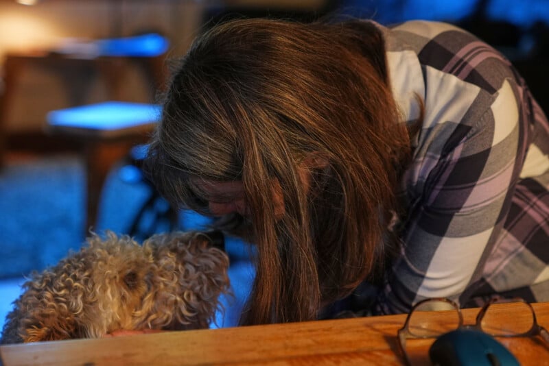 Junto a una mesa de madera, una mujer de largo cabello castaño y camisa a cuadros toca suavemente su frente con un caniche, creando un momento tierno en la habitación. En primer plano, sobre la mesa, hay gafas y un ratón de ordenador.