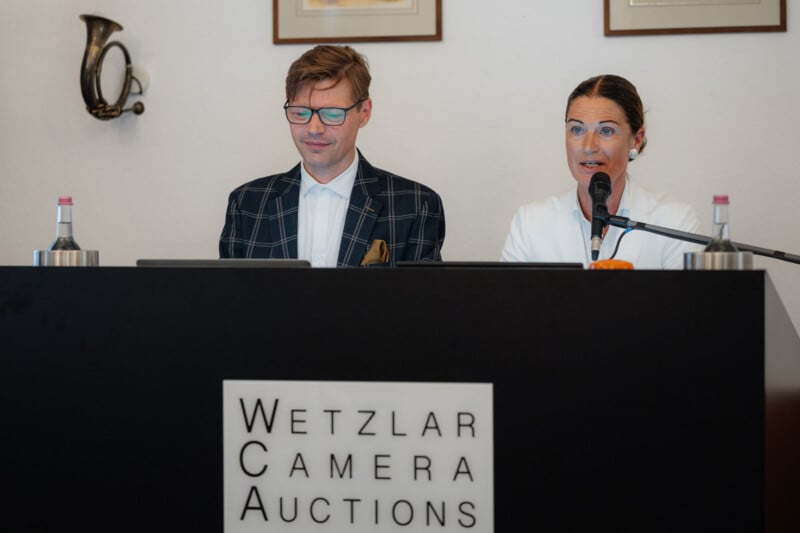 A man and a woman sit at a desk with laptops and microphones at a Wetzlar Camera Auctions event. The woman is speaking, while the man listens. Bottles of water and framed pictures are visible in the background.