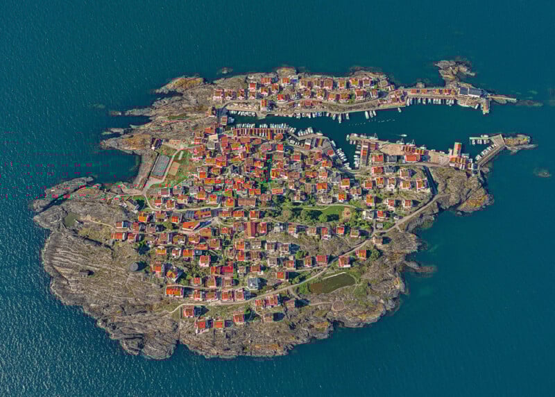 Vista aérea de una pequeña isla rocosa rodeada de aguas azules, salpicada de casas coloridas, tejados rojos, manchas verdes y muelles para embarcaciones amarradas.