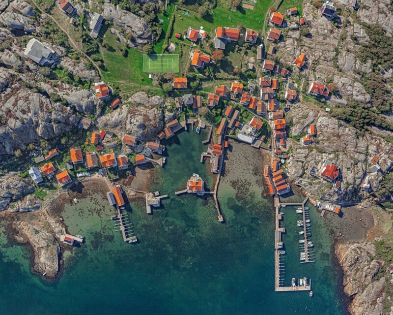 Vista aérea de un pueblo costero con casas con techos rojos agrupadas en un terreno rocoso, varios muelles que se extienden hasta el puerto verde azulado y una cancha de tenis cerca de la parte superior central de la imagen.