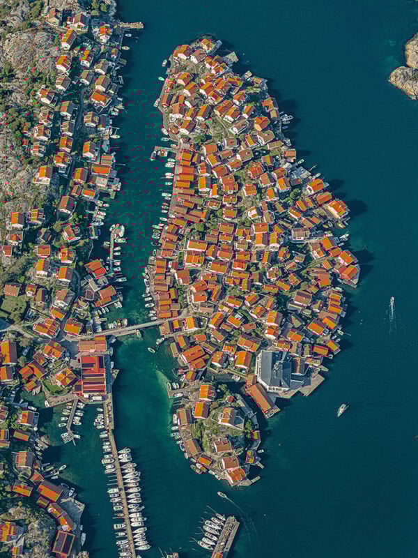Una vista de pájaro de una pequeña ciudad isleña con edificios densamente poblados con techos rojos, rodeada de agua azul y muelles con barcos amarrados. Un puente conecta la isla con el continente y varios barcos navegan cerca.