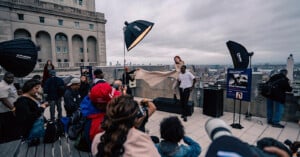 A rooftop photoshoot scene with a crowd watching as a photographer captures two models, one standing and one tossing fabric, against a cityscape. Large lights and camera equipment are set up around them.