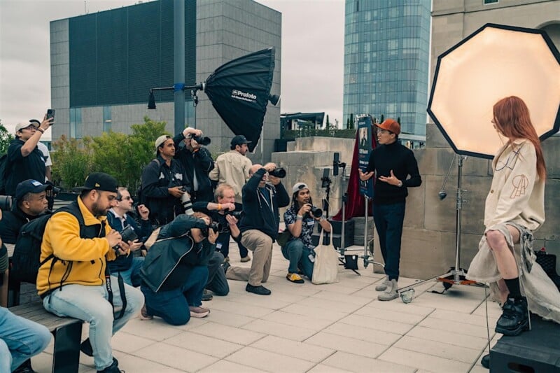 Un grupo de fotógrafos toma una fotografía de una mujer con largo cabello rojo posando frente a una gran caja de luz en una azotea, mientras un hombre está junto a ella dando instrucciones. Al fondo se ven claramente los edificios de la ciudad.