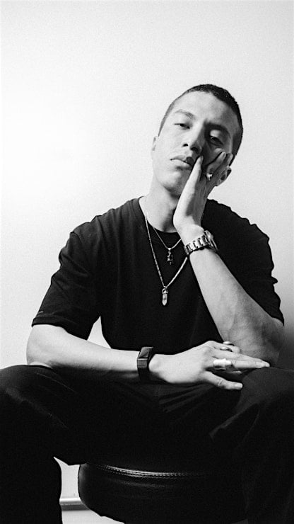 Un hombre está sentado en un taburete con un fondo liso, sujetándose la cara con una mano y mirando a la cámara. Llevan camisetas, collares, relojes y anillos oscuros y tienen expresiones neutras o pensativas.