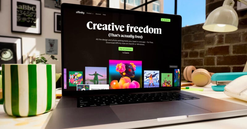 Computadora portátil sobre la mesa que muestra el sitio web de Affinity con el siguiente mensaje "Libertad creativa (en realidad gratis)" en la pantalla. Sobre la mesa hay una taza a rayas verdes, auriculares y varios materiales de arte.