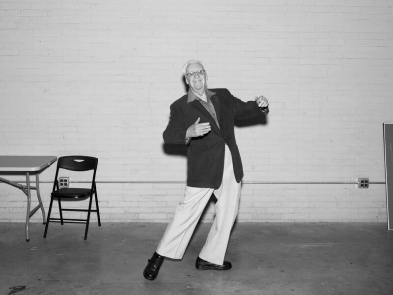 An elderly man wearing a blazer and light pants poses playfully in front of a white brick wall, near a folded chair and a table in an empty room.