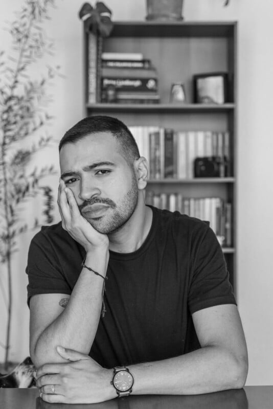 Black and white photo of a man with short hair and a beard, resting his face on his hand and looking at the camera, seated in front of a bookshelf filled with books and small decor items.