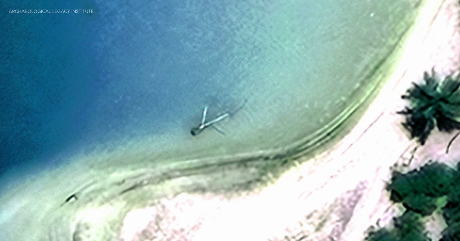 Aerial view of a coastline with clear blue water, sandy shore, and green vegetation. A partly submerged long structure or object extends from the shore into the water.