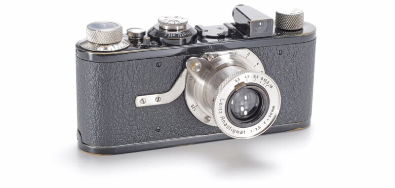 A vintage black and silver Leica camera with a textured body, prominent lens, and various dials on top, displayed on a white background.