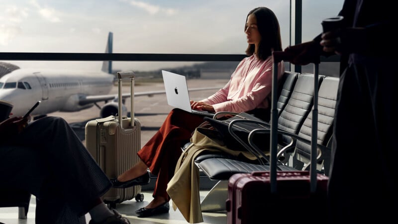 A woman sits in an airport terminal using a laptop on her lap, surrounded by suitcases. Large windows reveal an airplane parked outside on the runway. Other travelers are nearby.