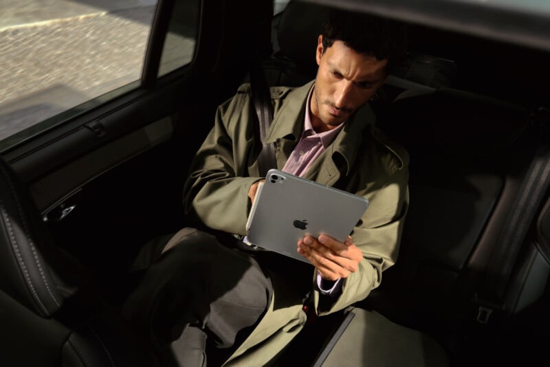 A man wearing a trench coat sits in the back seat of a car, looking intently at an iPad he is holding with both hands. Sunlight streams through the window, highlighting his focused expression.