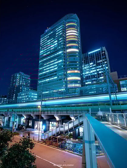 A cityscape at night with illuminated modern skyscrapers, a train moving quickly on elevated tracks, and streetlights casting a blue glow on the surrounding area below.