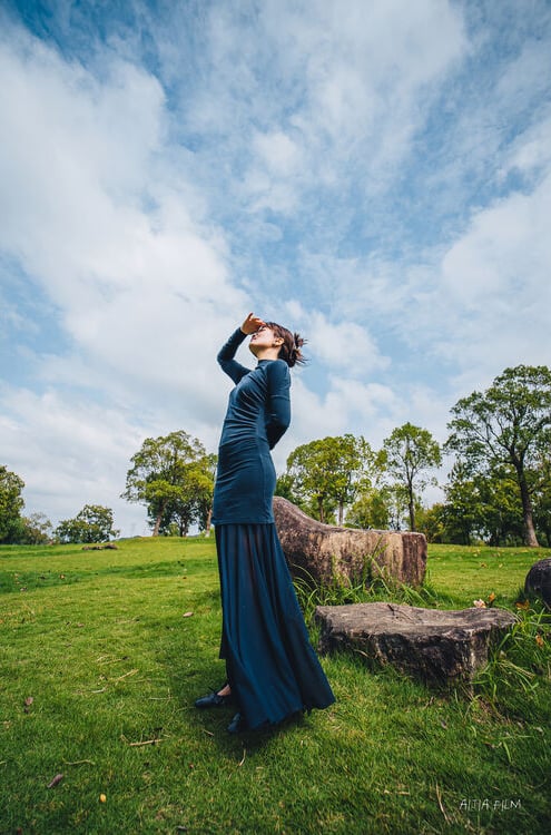 A woman in a long dark blue dress stands on grass beside large rocks, looking up with one hand shielding her eyes. Green trees and a partly cloudy sky fill the background.