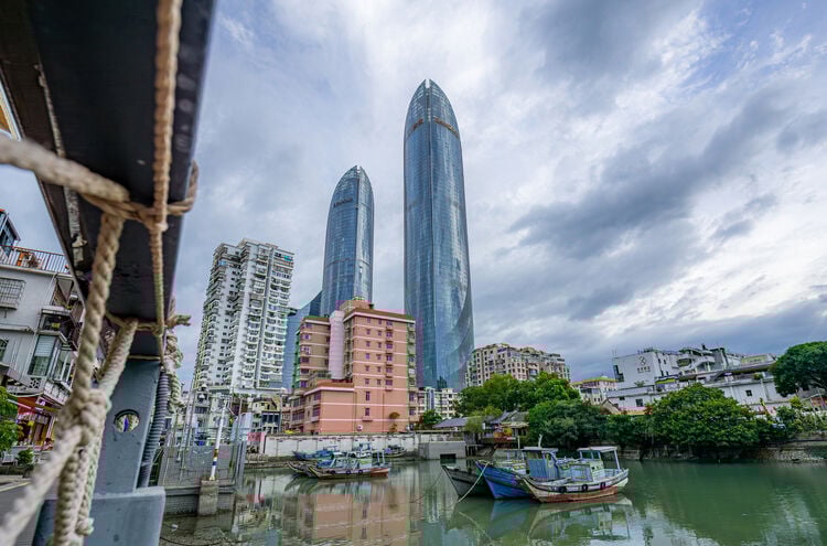 Two modern glass skyscrapers rise above older buildings and docked boats along a calm riverside under a cloudy sky in an urban cityscape.