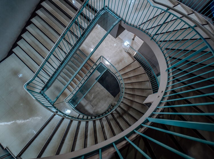 A spiral staircase with teal railings viewed from above, creating a geometric pattern as it curves around a central open space. The steps and floor reflect light, enhancing the modern architectural design.