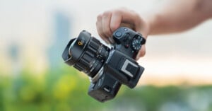 A person holds a black digital camera with a large lens in their right hand; the background is blurred with hints of green and blue.
