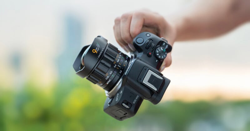 A person holds a black digital camera with a large lens in their right hand; the background is blurred with hints of green and blue.
