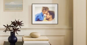 A framed photo of a man and a young girl affectionately touching foreheads hangs on a beige wall above a table with a plant, a round stone object, and a drawer. Another landscape painting is seen on the wall.