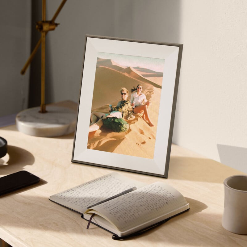 Una fotografía enmarcada de dos personas sentadas en una duna sobre una mesa de madera clara con un cuaderno abierto, un bolígrafo y una lámpara cerca. El sol proyecta suaves sombras sobre la escena.