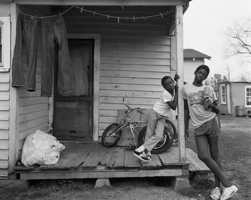 Two children stand on a worn wooden porch; one leans on a post while the other stands with crossed legs, holding something. A bicycle is behind them, with laundry hanging and a plastic bag pile nearby. The house looks weathered.