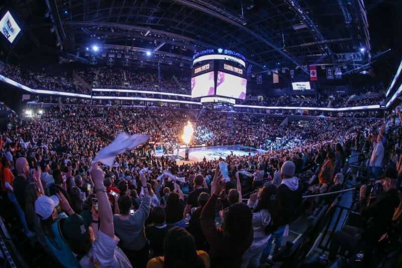 A packed basketball arena filled with cheering fans, many waving white towels, as flames shoot up from the court during a pre-game or halftime show. The scoreboard hangs above the court, and the atmosphere is energetic.