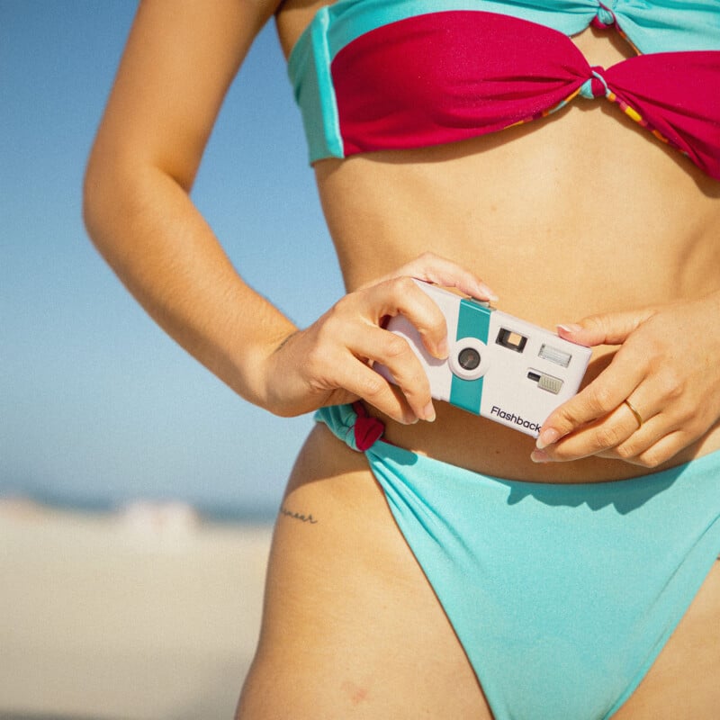 A person in a turquoise and pink bikini holds a white disposable camera at their waist while standing on a sunny beach with blue sky in the background.