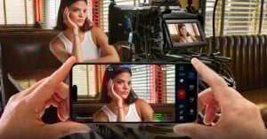 A woman sits in a diner booth, resting her head on her hand, as she is filmed by a camera. A person holds up a smartphone displaying a live recording of the woman in the foreground.