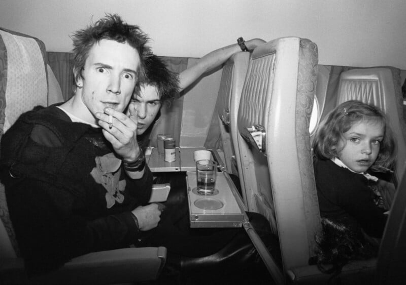 Two young men with punk hairstyles and dark clothes sit in airplane seats, one looking surprised at the camera. A young girl sits beside them, looking toward the camera with a neutral expression. Airplane trays hold drinks.