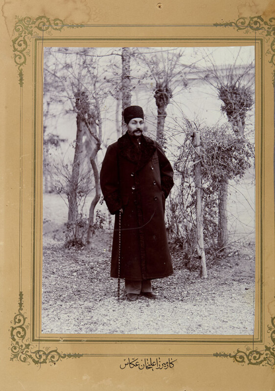 Un hombre vestido con un abrigo largo y oscuro y un sombrero posa para una fotografía al aire libre cerca de árboles desnudos. La foto en tonos sepia presenta un borde decorativo y texto persa en la parte inferior.