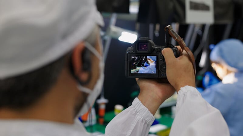 A person in protective clothing and a face mask is photographing a workplace scene with a camera, focusing on someone working at a desk, also dressed in protective attire.