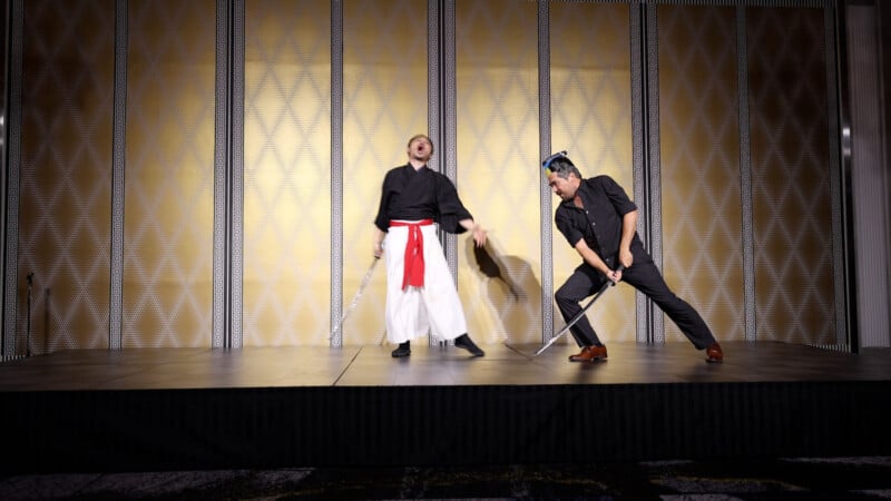 Two men perform a sword fight on a stage. The man on the left wears a black top with white pants and a red sash, standing with his mouth open. The man on the right wears black and holds his sword defensively.