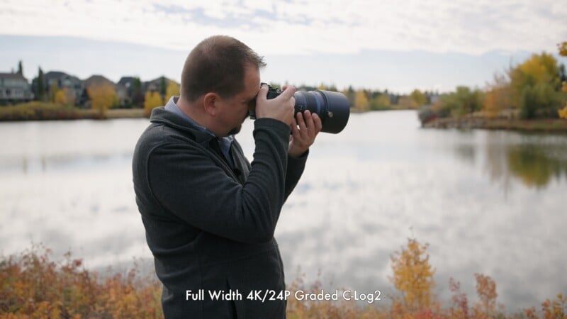 A man stands by a lake taking a photo with a camera, surrounded by autumn foliage and houses in the background. Text at the bottom reads, "Full Width 4K/24P Graded C-Log2.