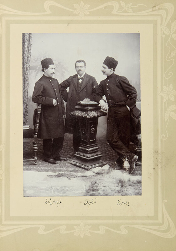 Tres hombres con ropas y sombreros tradicionales oscuros posan en un estudio antiguo, de pie alrededor de un pedestal decorativo. Los dos hombres se apoyan el uno contra el otro casualmente, mientras que el hombre del medio se mantiene erguido. El fondo y el suelo están profusamente decorados.