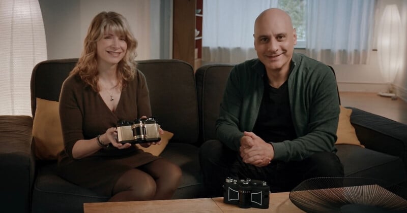 Una mujer y un hombre se sientan en un sofá en una cómoda sala de estar y sonríen a la cámara. La mujer sostenía un pequeño dispositivo robótico y otro dispositivo idéntico estaba colocado sobre la mesa frente a ellos.