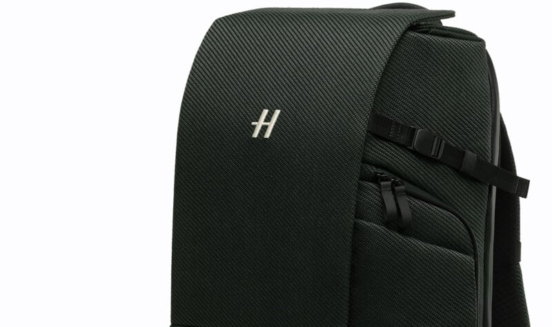 A close-up of a dark green textured backpack with a flap, featuring a silver "H" logo and a zippered front pocket with a black adjustable strap on the side.