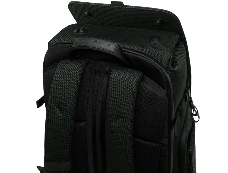A close-up of a dark green backpack with padded shoulder straps and an open top flap, revealing a black label. The texture of the fabric and zipper details are visible.