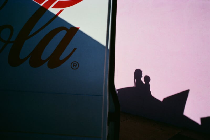 Shadow of an adult and a child on a pink wall, partially covered by the shadow of a van with a Coca-Cola logo. Bright sunlight creates contrasting colors and sharp lines.