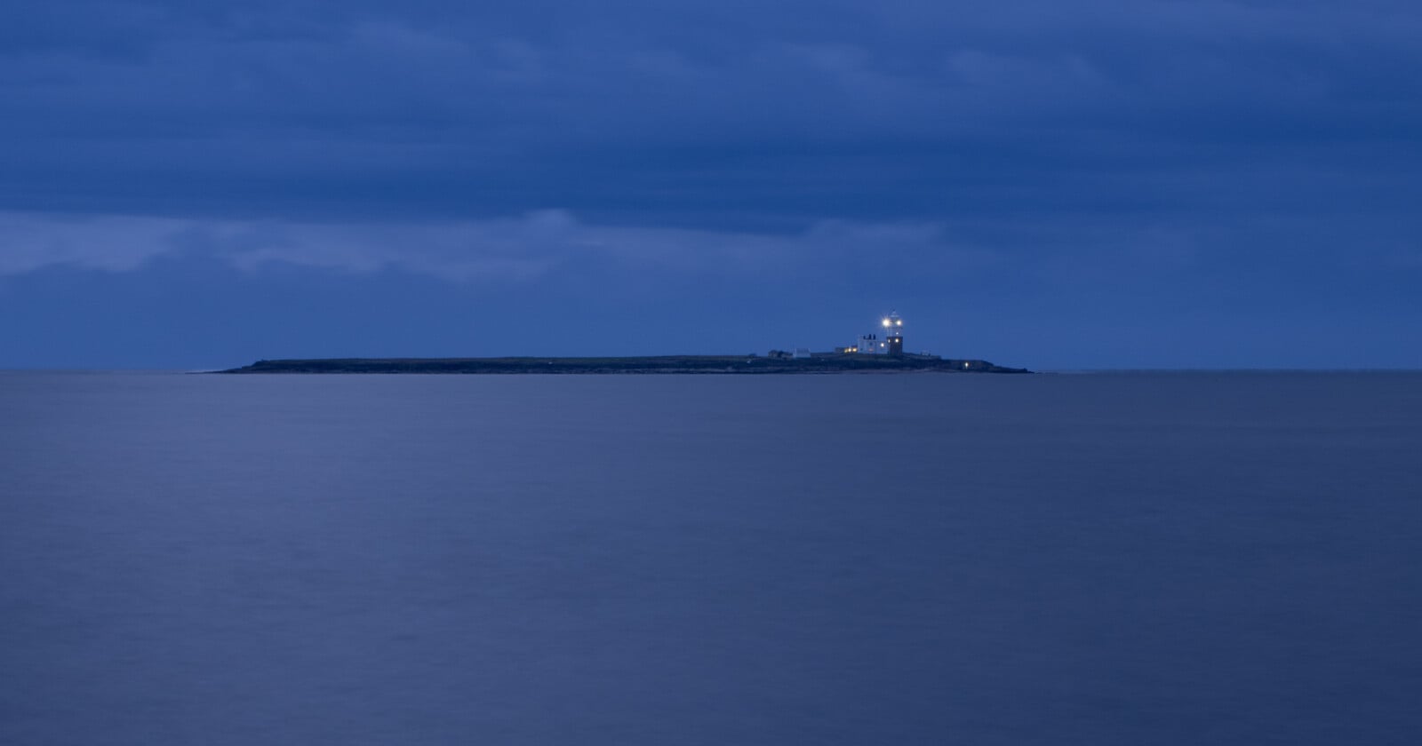 Coquet island with a lighthouse illuminated at dusk, surrounded by calm, blue ocean water under a cloudy sky.