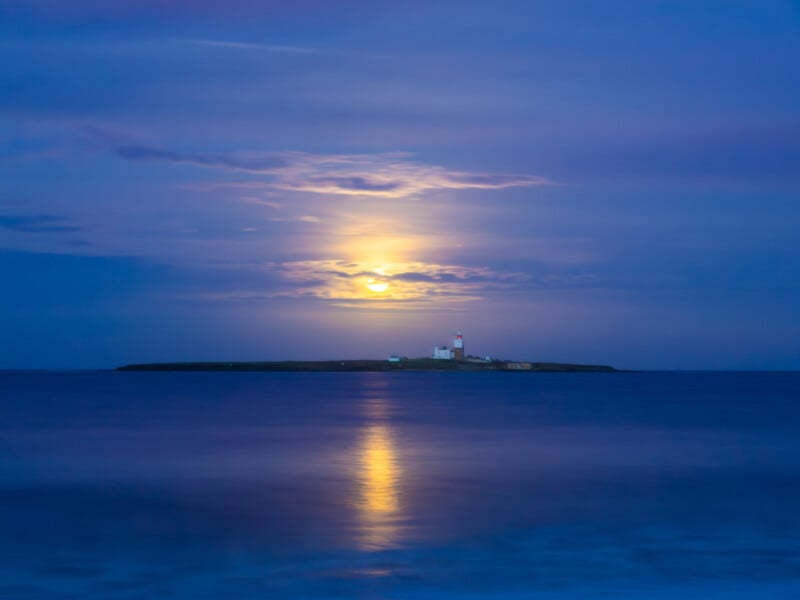 Una pequeña isla con un faro se encuentra en tranquilas aguas azules bajo un cielo nublado, con una brillante luna llena reflejada en la superficie.