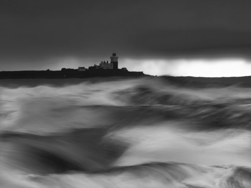 Una fotografía en blanco y negro de olas borrosas rompiendo en primer plano, un cielo oscuro y espectacular al fondo y la silueta de un faro en una pequeña isla en la distancia.
