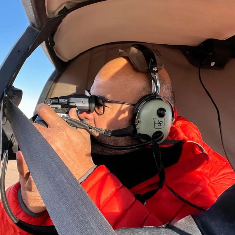 A person in a red jacket and headset is seated inside an aircraft, looking out the window while holding a camera, ready to take a photo. Sunlight illuminates their face and the plane cabin interior.